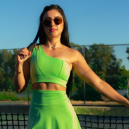 Fitness model posing with a racquet on a tennis court wearing a green apple, mini skort matching set from vibras activewear.